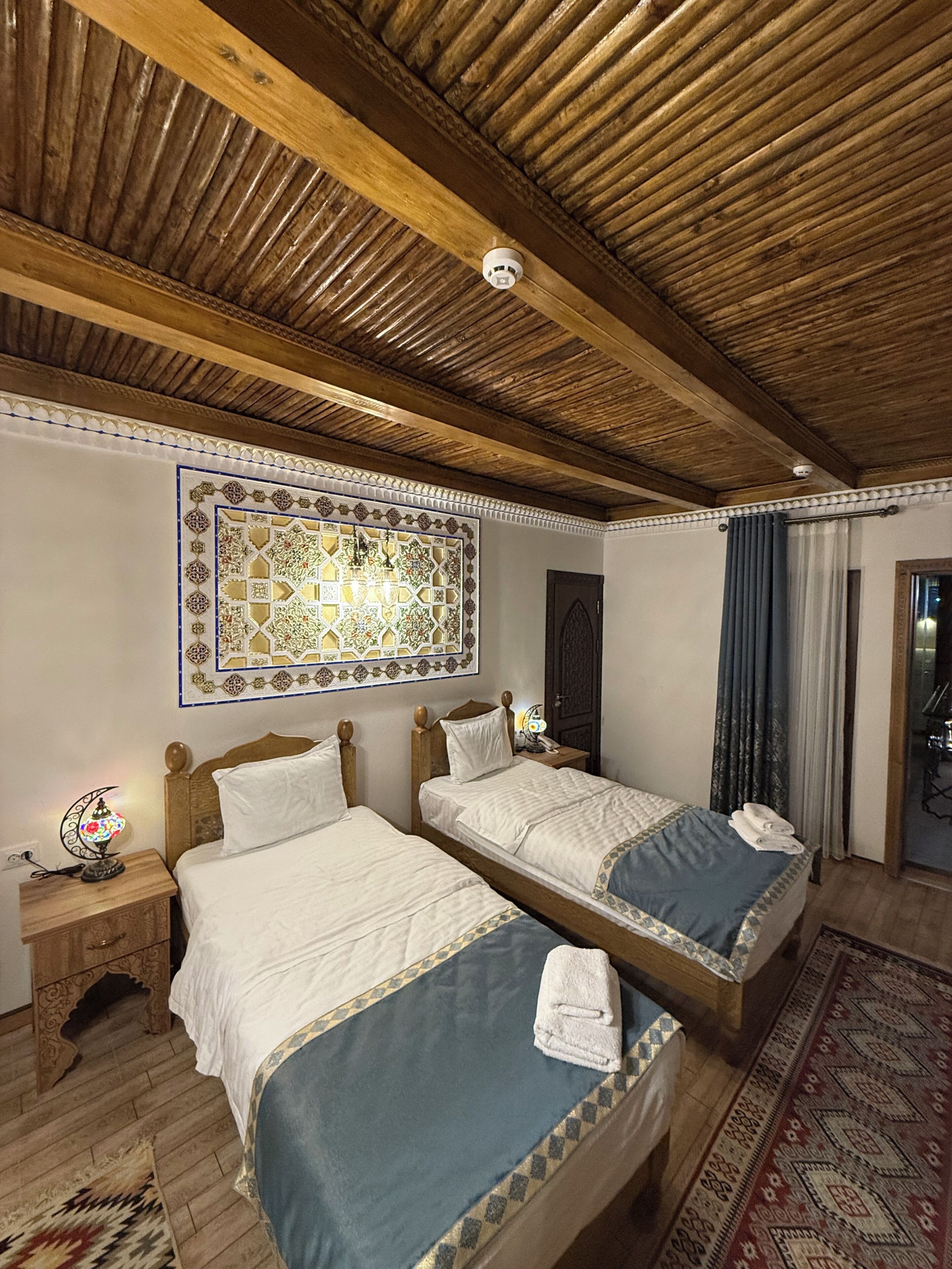 A room with two wooden beds, a patterned wall decoration, a nightstand with a lamp, and a door leading to another room.