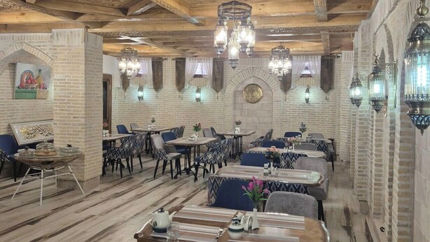 A restaurant with a high ceiling, patterned walls, and traditional chandeliers. Tables are set with striped tablecloths and patterned chairs. - Mehrob Hotel (Bukhara)