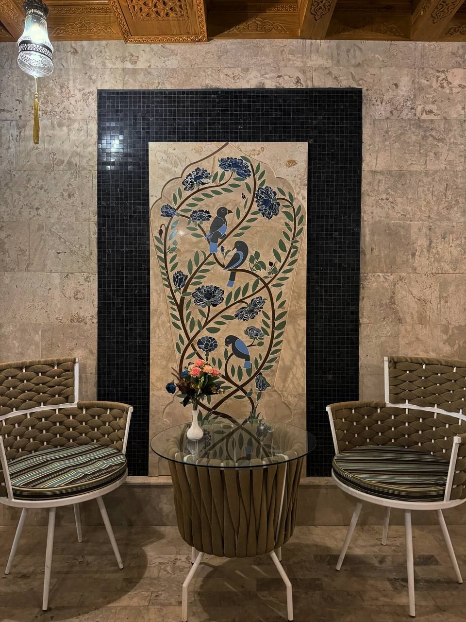 Um quarto com mesa de vidro, duas cadeiras estofadas, um objeto decorativo na parede e um lustre.