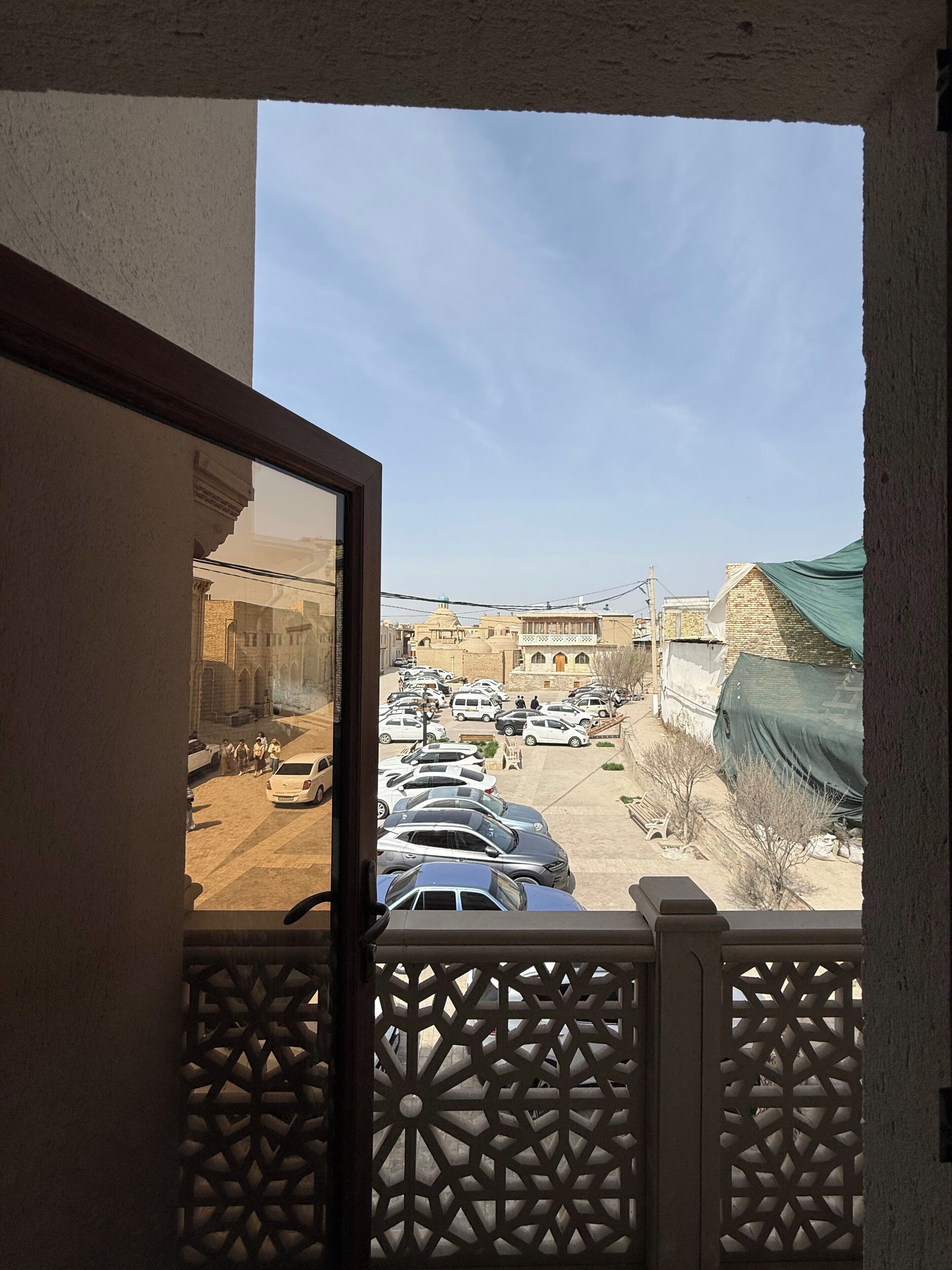 Une vue depuis un balcon doté d’une balustrade décorée, donnant sur un parking où se trouvent plusieurs voitures, avec des bâtiments en arrière-plan.