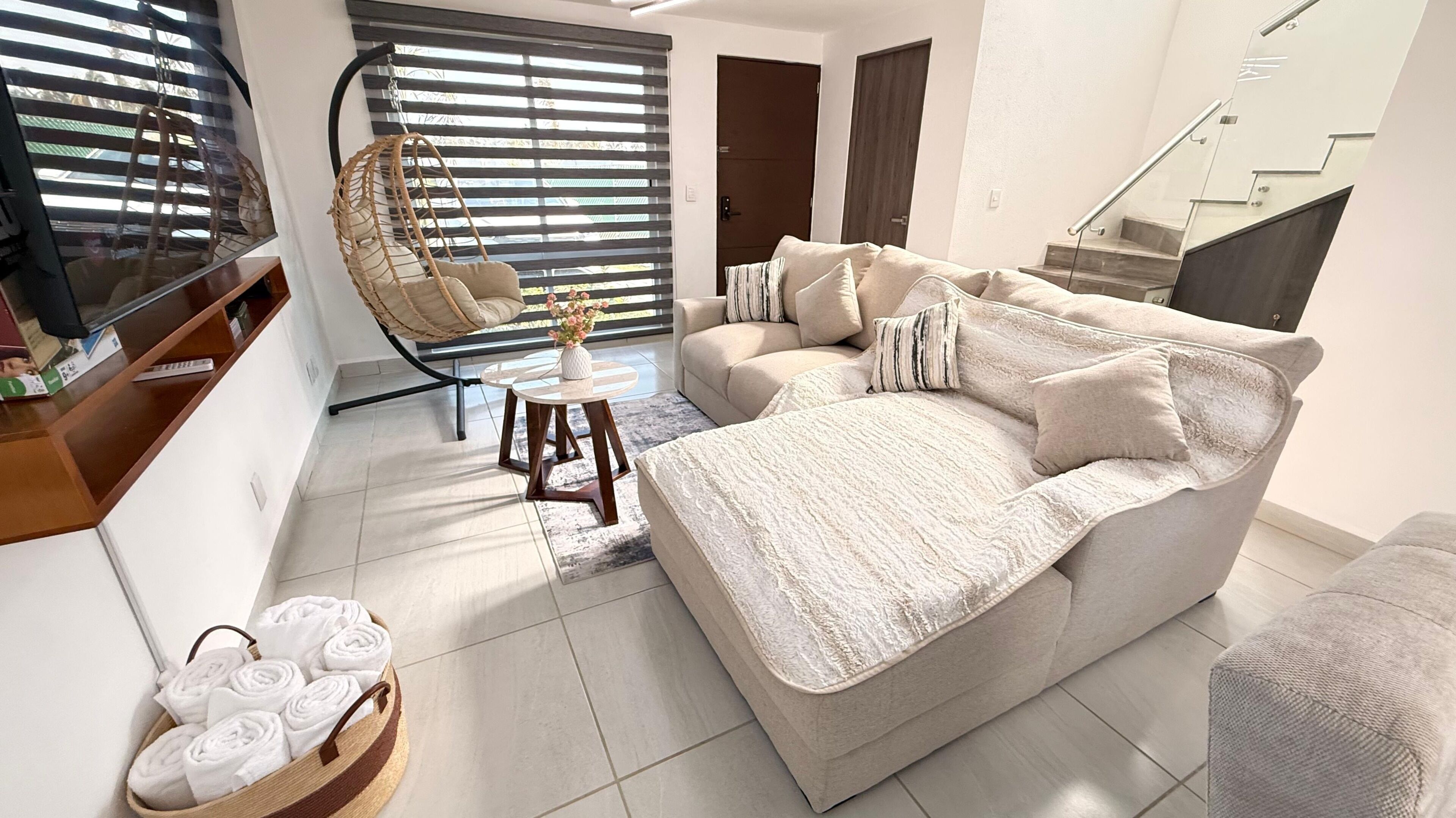 Una sala de estar moderna con un sofá, una silla colgante, una mesita con un jarrón y una cesta de toallas.