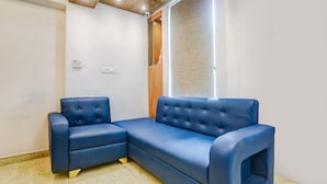 A room with a blue sectional sofa, a small table, a wooden cabinet, and a window with blinds. - Deluxehotel Riash (Prayagraj)