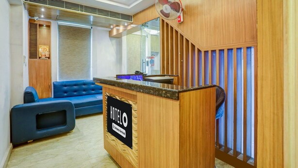 A hotel reception area with a wooden reception desk, a blue sofa, and a fan. - Deluxehotel Riash (Prayagraj)