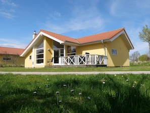 Ein gelbes Gebäude mit rotem Dach, einer Veranda mit weißen Geländern und einer Blumenwiese davor.