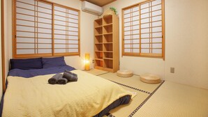 A traditional Japanese room with tatami flooring, a bed with a yellow patterned blanket, and a wooden shelf with a plant. - Lucky Higashi Mizue (Tokyo)
