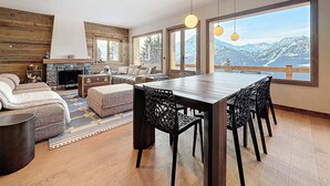 Uma sala de estar moderna com lareira, área de jantar com mesa grande e cadeiras, e amplas janelas com vista para as montanhas.