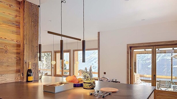 Uma cozinha moderna com armários de madeira, uma ilha grande, luminárias pendentes e vista para montanhas nevadas através de amplas janelas.