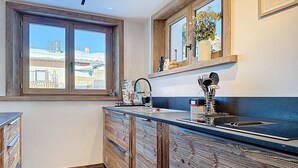 Uma cozinha com armários de madeira, bancada preta, janela com vista para prédios e um vaso de flores no parapeito.