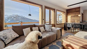 Uma sala de estar moderna com sofá, mesa de jantar e cadeiras, grandes janelas com vista para as montanhas, e uma parede de destaque em madeira.