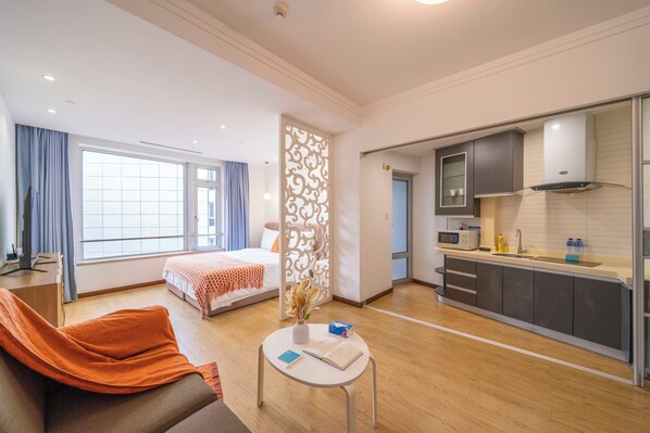 Ein modernes Hotelzimmer mit einem Bett, einer Couch, einem kleinen Tisch mit Buch und Vase, einer Küchenzeile mit Schränken und einer Mikrowelle sowie einem großen Fenster mit Vorhängen.