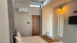 A hotel room with a bed, a wall-mounted air conditioner, a door, a towel on the bed, a wall-mounted TV, and a window. - Hotel Mojito (Vyara)