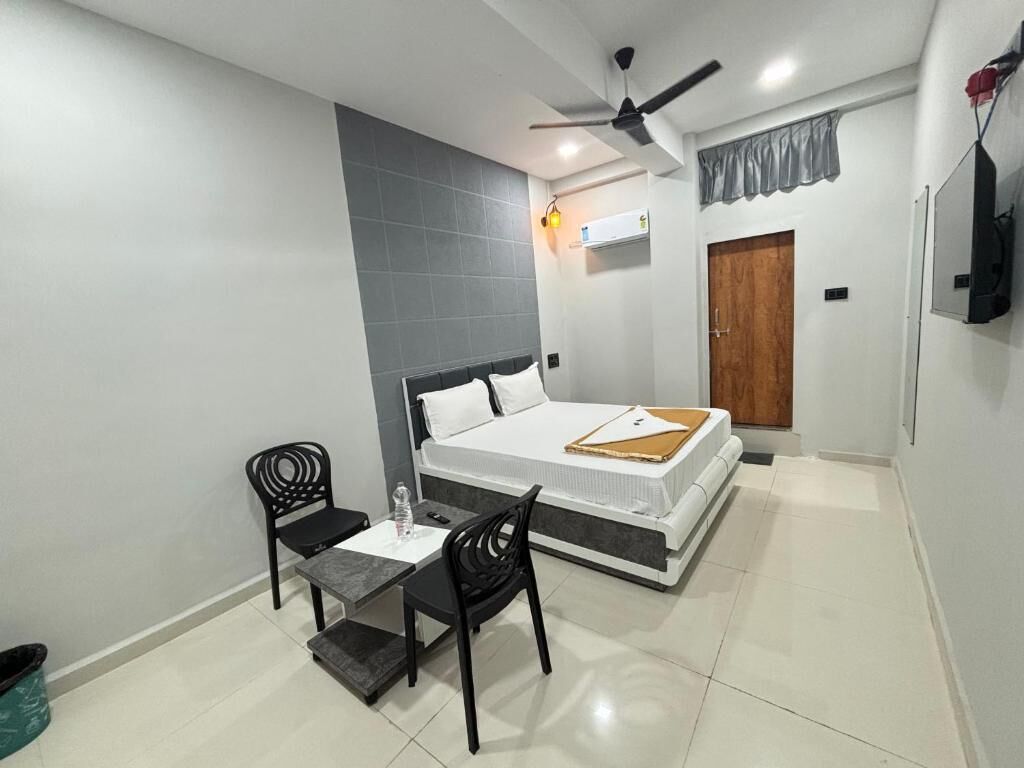 A bedroom with a bed, a desk, two chairs, a ceiling fan, an air conditioning unit, and a wooden door.