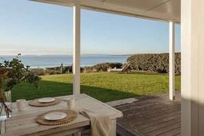 Eine überdachte Terrasse mit Tisch für zwei Personen, mit Blick auf eine Rasenfläche und das Meer.