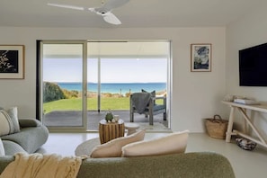 A modern living room with a large sliding glass door leading to a deck, a sofa, a small round table, and a television mounted on the wall. - 'Kindred' Absolute Beachfront Retreat (Callala Beach)