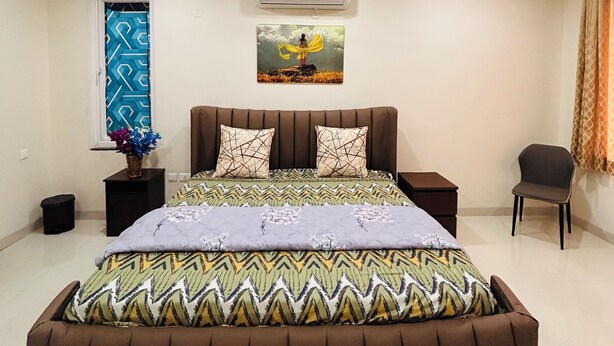 A bedroom with a bed, a ceiling fan, an air conditioner, a window with a geometric pattern, a wall-mounted picture, a bedside table, and a chair. - Luxury 4BHK Villa Near Airport, Hyderabad Family + Work Friendly (Hyderabad)