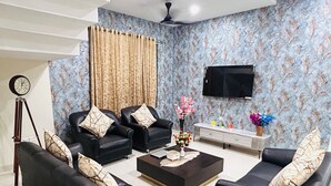 A living room with two black leather sofas, a coffee table, a TV, and a wall-mounted clock. - Luxury 4BHK Villa Near Airport, Hyderabad Family + Work Friendly (Hyderabad)