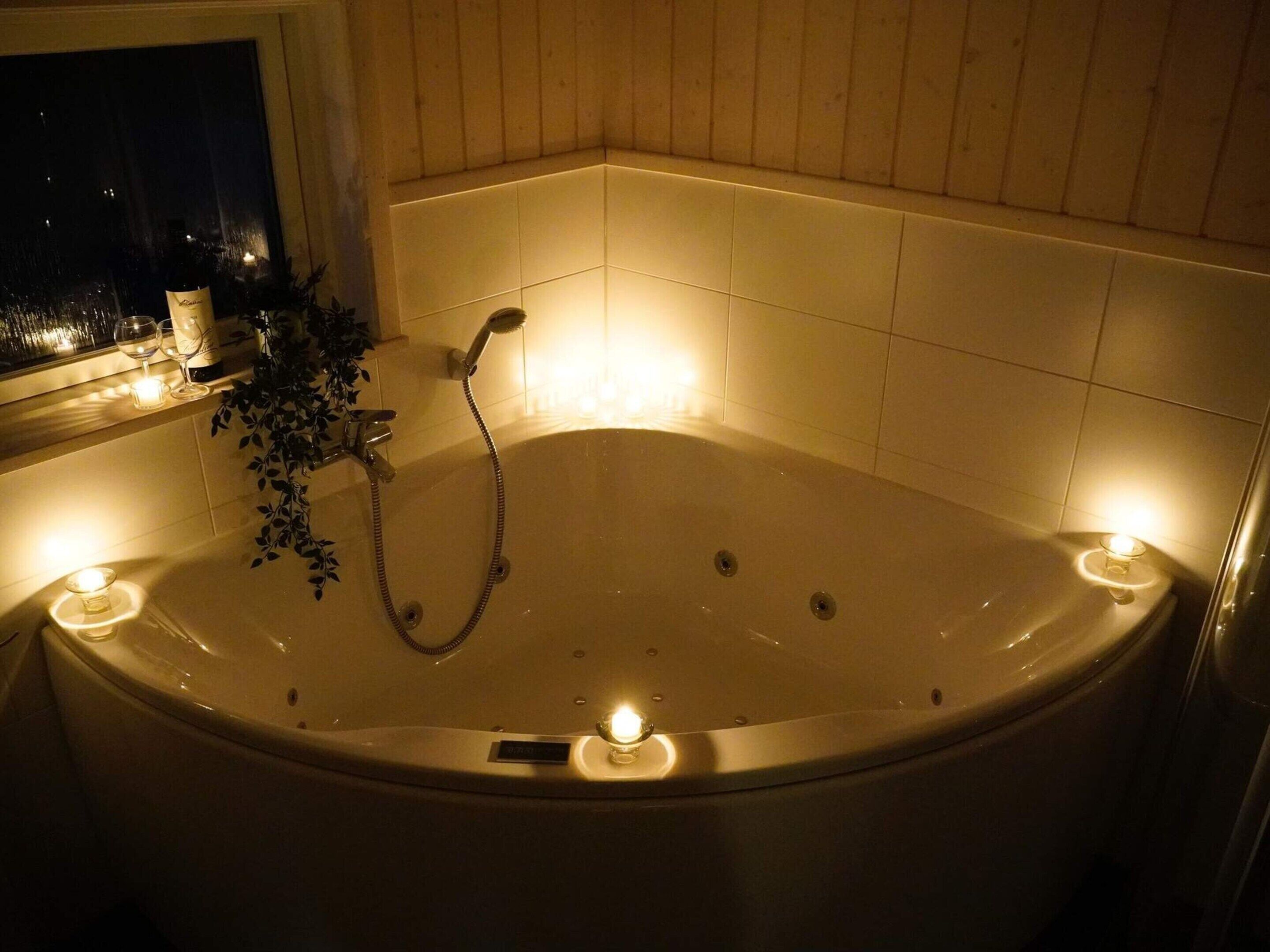 A large, oval-shaped bathtub with a showerhead, surrounded by candles, in a room with wooden paneling and a window.