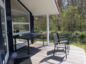 Ein überdachter Außenbereich mit Tisch und Stühlen, einer Holzterrasse und Blick auf die umliegende Natur.