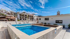 Ein rechteckiger Pool mit Fliesen umgeben, umrahmt von einem weißen Gebäude mit einem ebenfalls gefliesten Dach und einem Balkon.