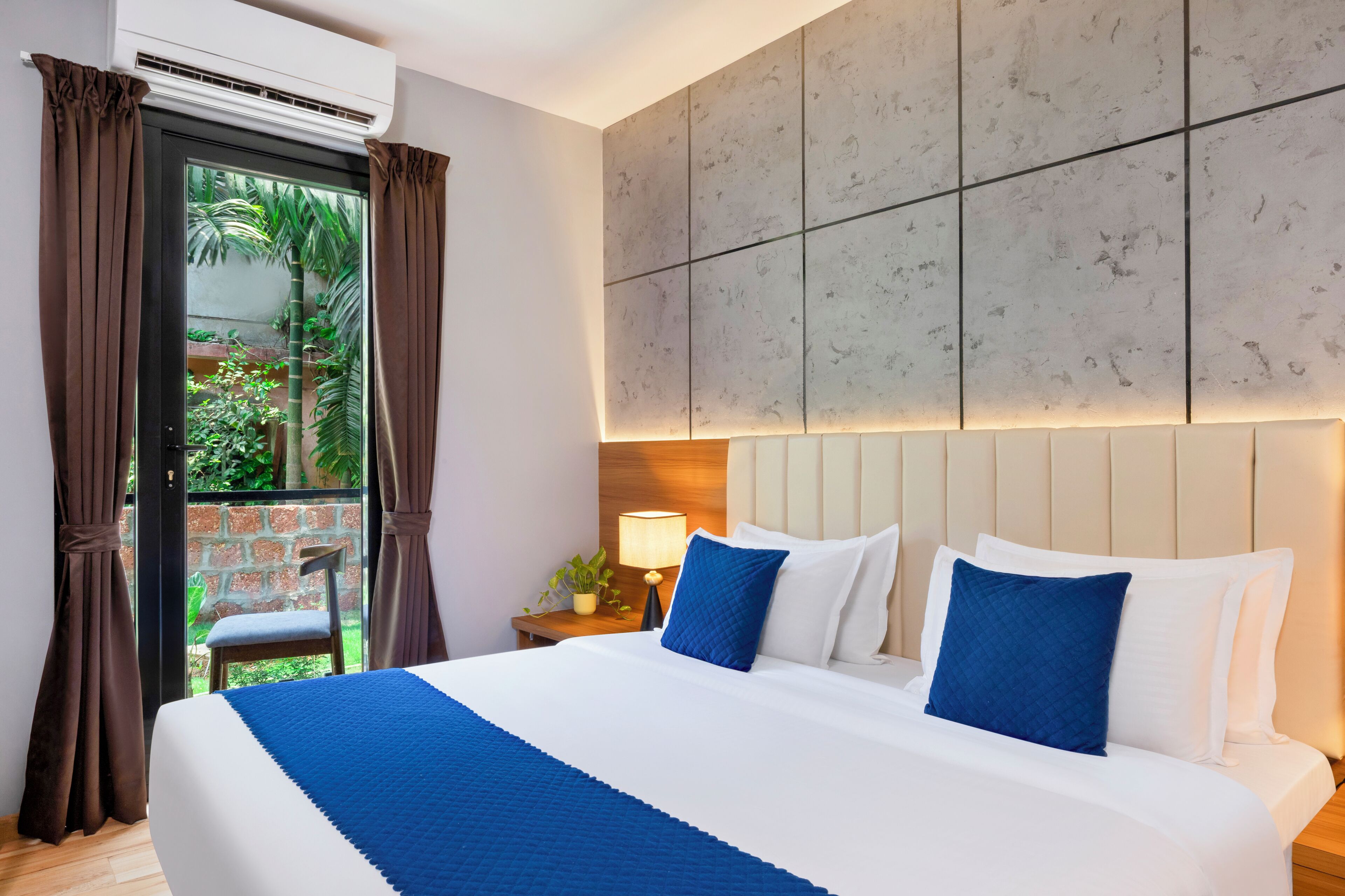 A hotel room with a bed, a nightstand, a lamp, and a view of greenery outside.