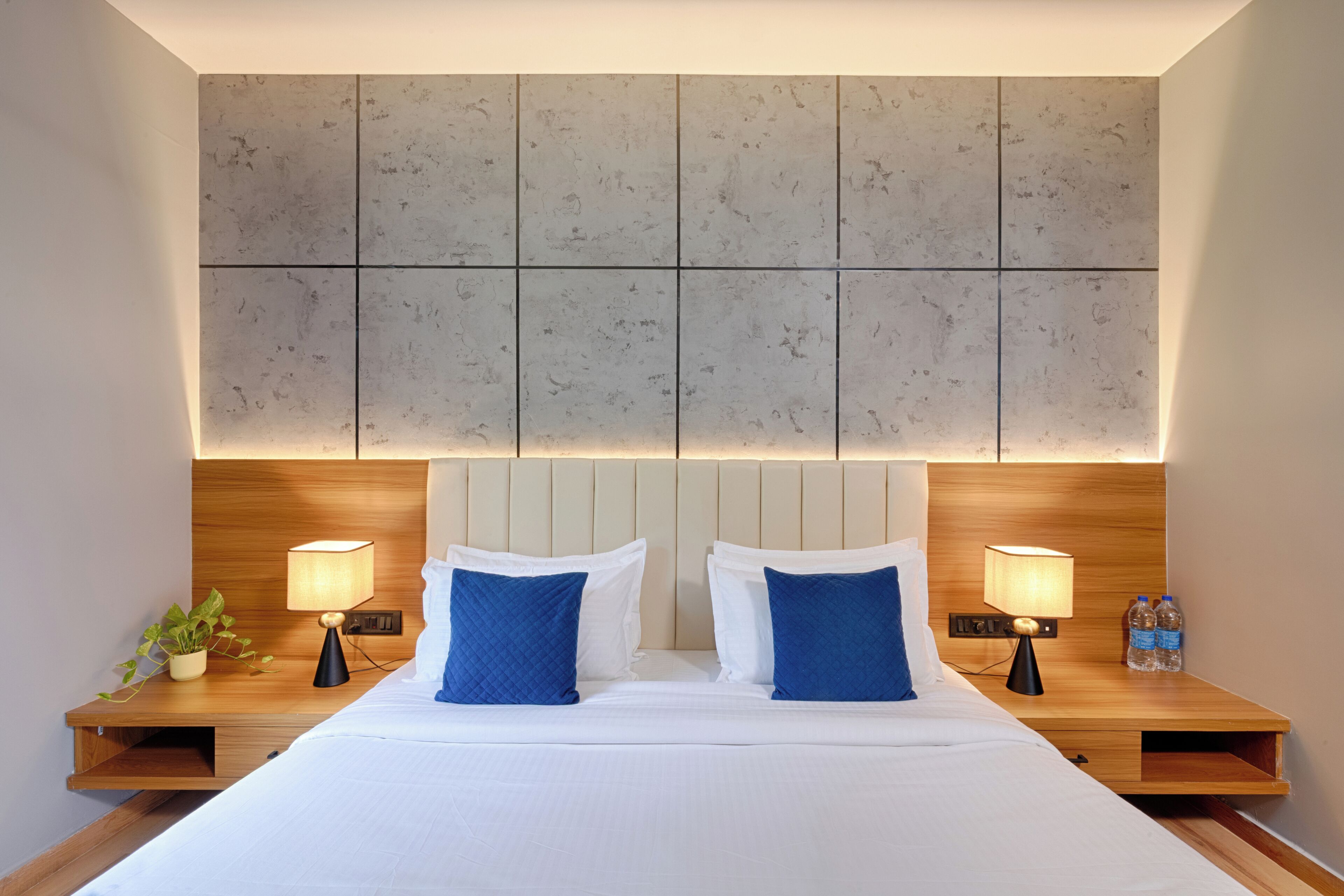Habitación de hotel con una cama grande, dos mesitas de noche, dos lámparas y un cabecero fijado a la pared.