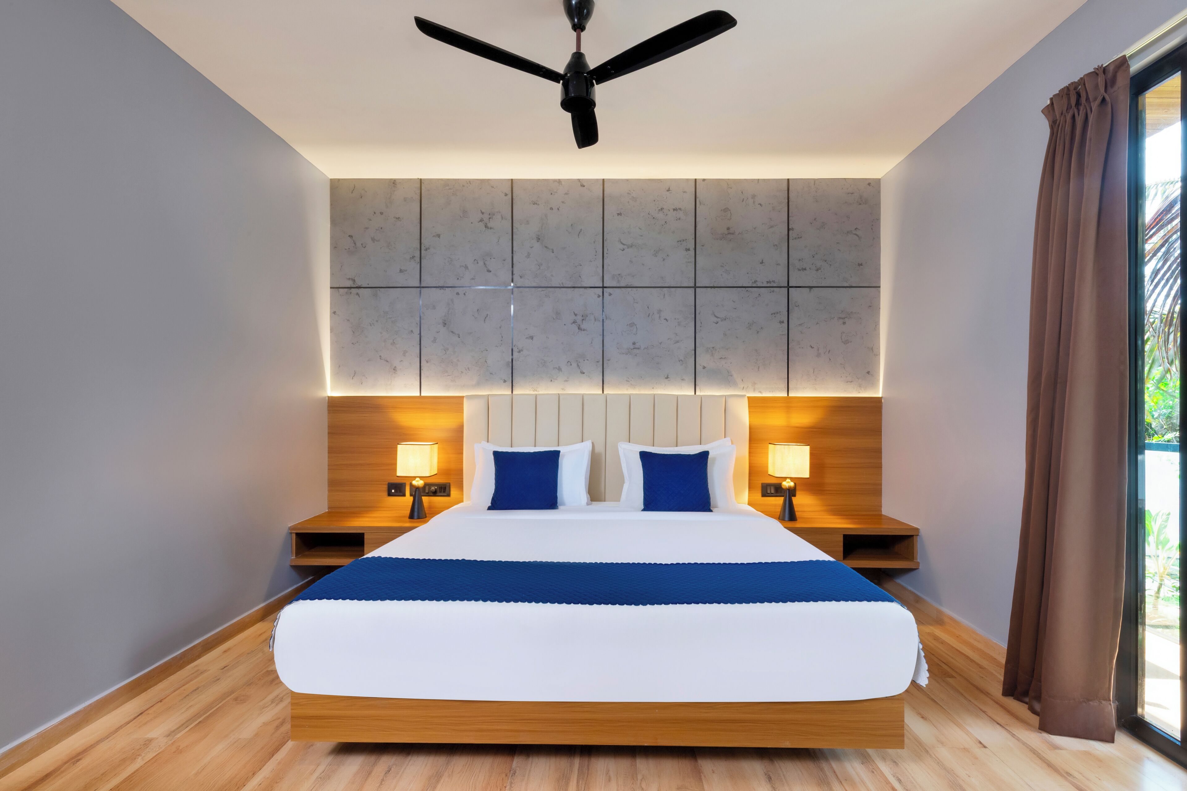 Une chambre d’hôtel moderne avec un grand lit, deux tables de chevet avec des lampes, un ventilateur de plafond et une vue sur l’extérieur à travers une fenêtre avec des rideaux.