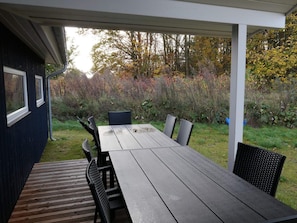 Un portico coperto con tavolo e sedie in legno, immerso in un giardino con alberi e arbusti.