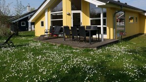 Um edifício amarelo com alpendre coberto, mesas ao ar livre e um gramado com flores brancas.