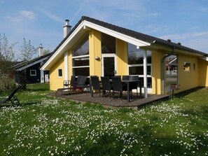 Ein gelbes Gebäude mit überdachter Terrasse, Außenbestuhlung und einem Rasenbereich mit weißen Blumen.