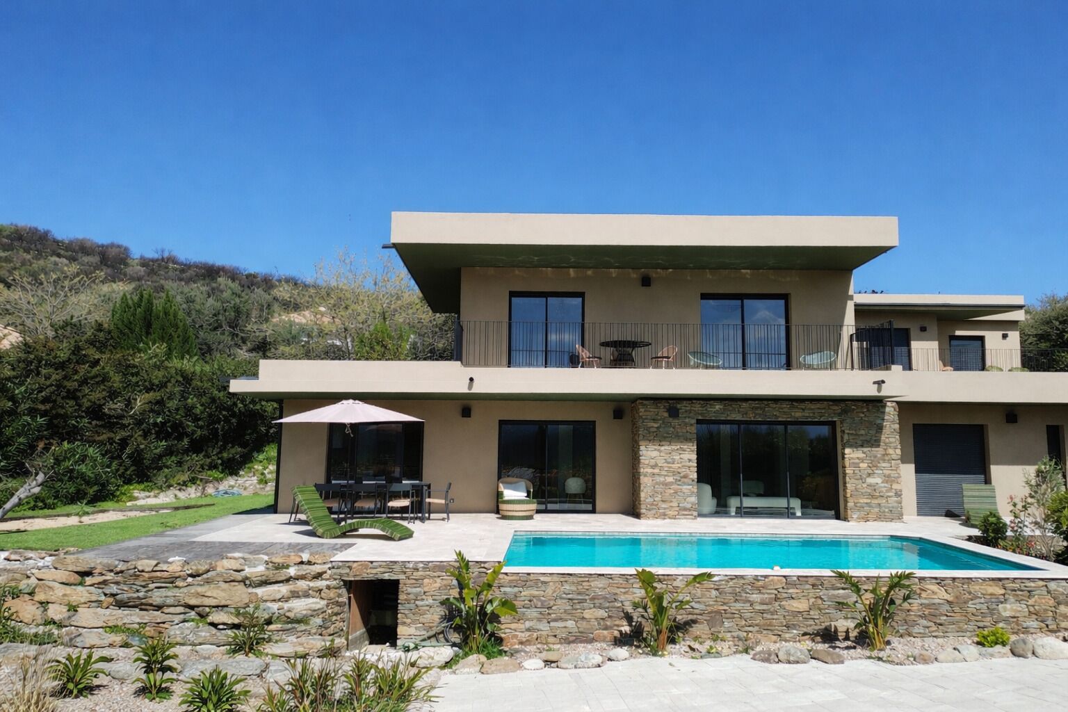 Une maison moderne à deux étages dotée d’une piscine, d’un aménagement paysager en pierre et d’un patio avec mobilier d’extérieur.