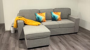 Een grijze bank met kleurrijke kussens, een gele plaid en een witte speaker op een houten vloer.