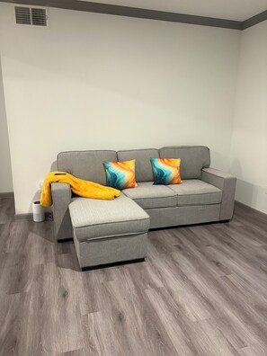 Een grijze bank met kleurrijke kussens, een gele plaid en een witte speaker op een houten vloer.
