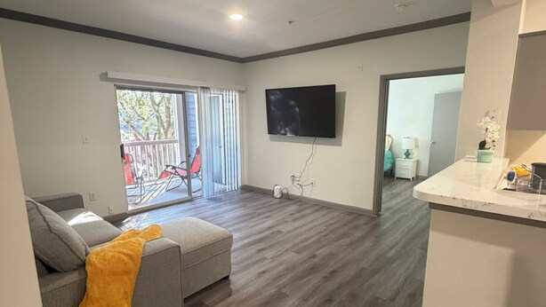 Een moderne woonkamer met een grijze bank, een gele plaid, een flatscreen televisie aan de wand en een houten vloer.