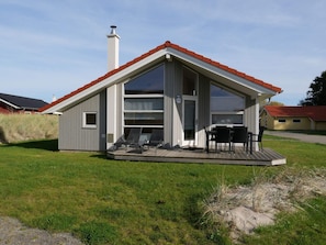 Uma casa moderna com telhado inclinado, chaminé, deck com cadeiras e uma janela grande.