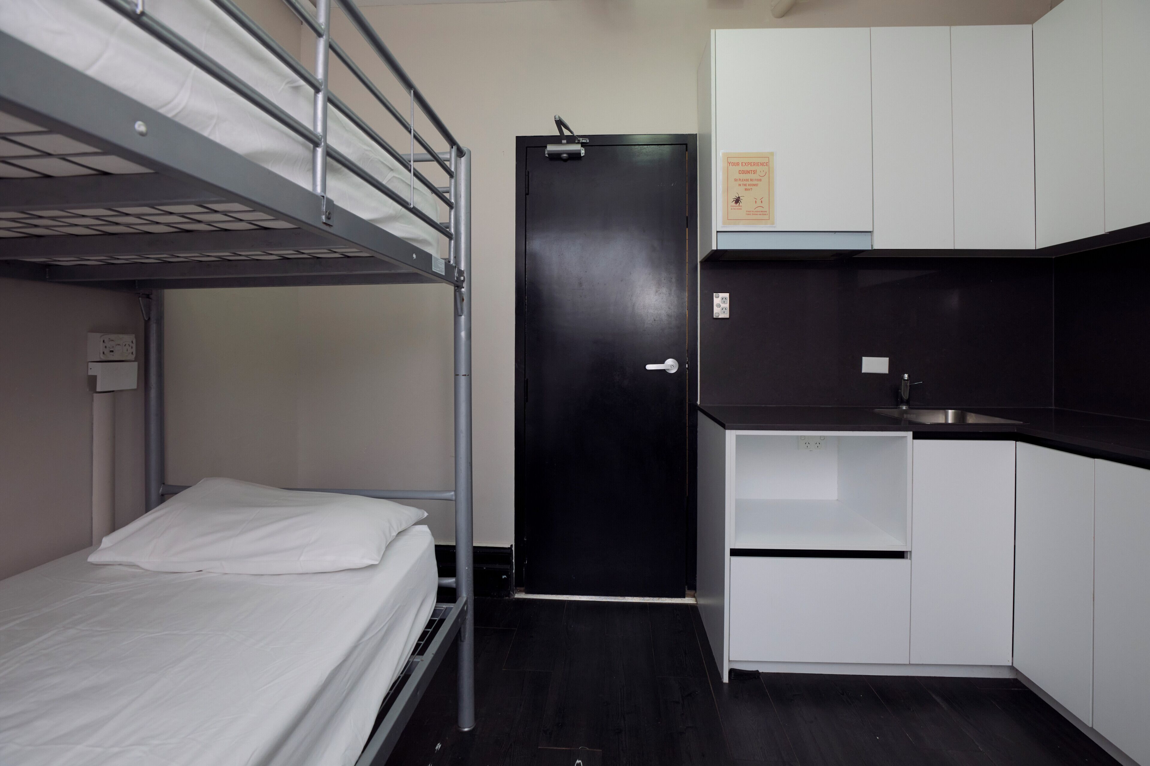 Een stapelbed, een zwarte deur, een keuken met witte kasten en een klein plankje.