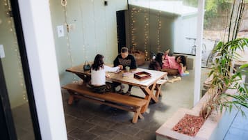 A group of people seated at a wooden table with benches, surrounded by string lights, in a room with a tiled floor and a large window.