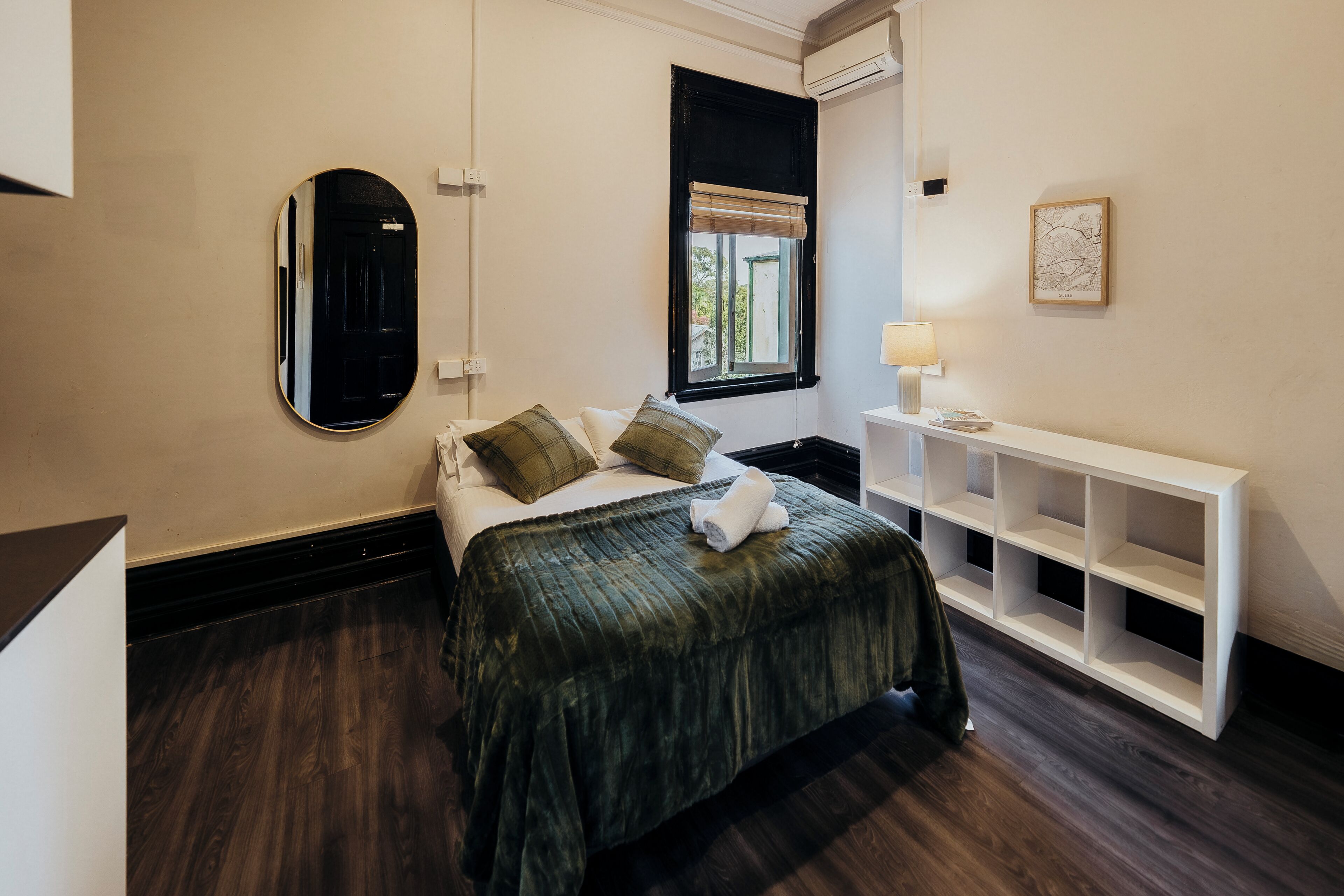 A hotel room with a bed, a bedside table, a lamp, a window with a view, and a wall-mounted air conditioner.