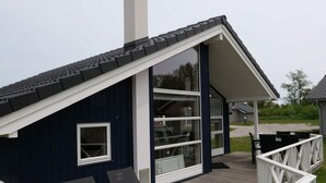 A small, dark blue building with a sloped roof, large windows, and a wooden deck with chairs. - 4 Star Holiday Home in Grossenbrode (Grossenbrode)