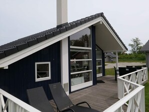 A small, dark blue building with a sloped roof, large windows, and a wooden deck with chairs. - 4 Star Holiday Home in Grossenbrode (Grossenbrode)