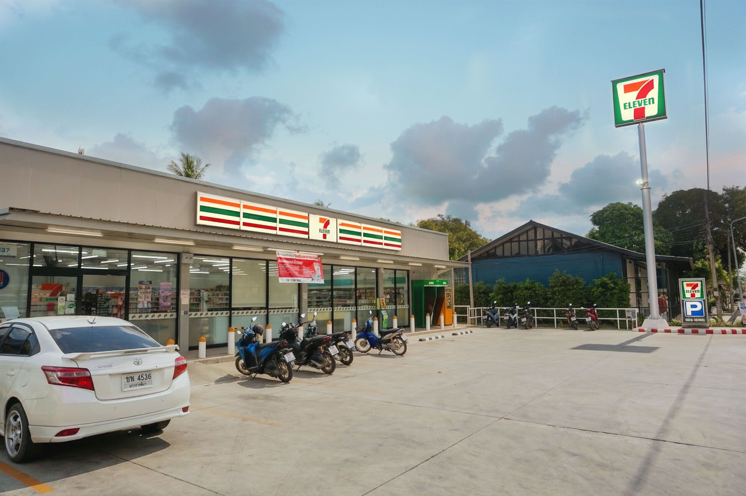 Una tienda 7-Eleven con un letrero de estacionamiento, un coche blanco y varias motocicletas estacionadas frente.
