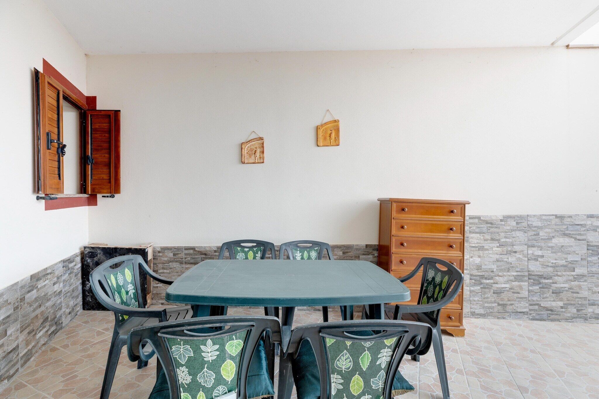 A dining area with a table and chairs, a wooden cabinet, and a wall with a window and two hanging decorations.