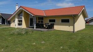 Uma casa amarela com telhado vermelho, um pátio com cadeiras e um céu azul límpido.