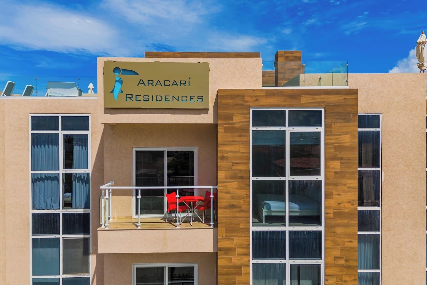 En modern byggnad med en skylt som visar texten ”Aracari Residences”. Den har flera fönster, en balkong med röda stolar och en träfasad.
