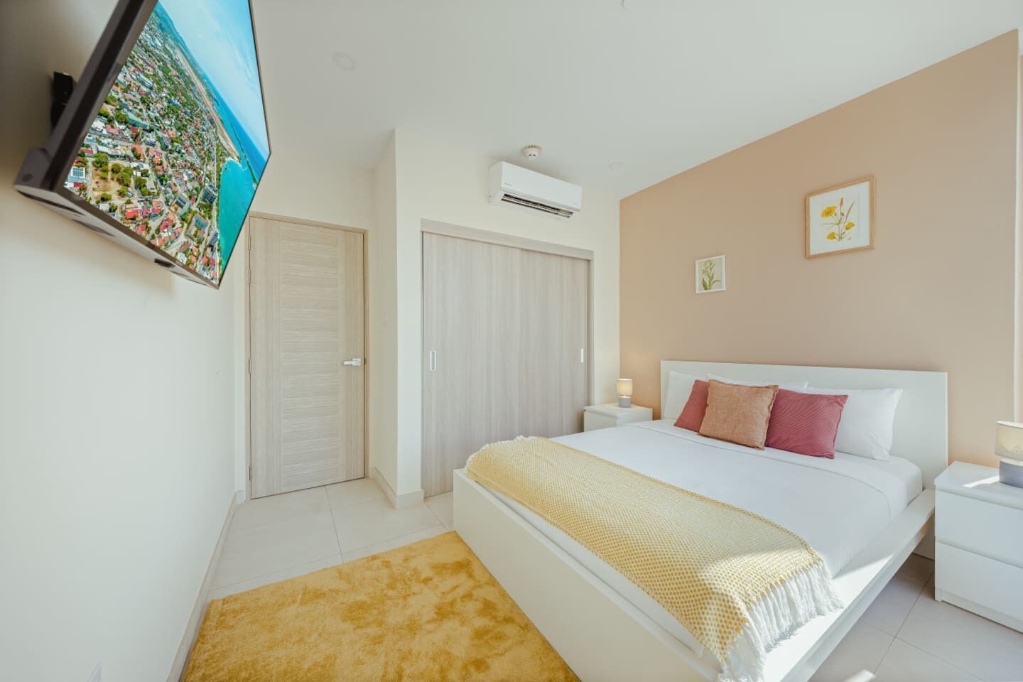 A bedroom with a bed, a TV mounted on the wall, an air conditioning unit, and a view of a cityscape.