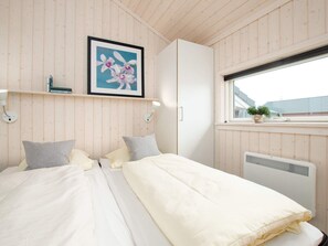 Ett sovrum med en säng, kuddar, en inramad tavla, en hylla med böcker, ett fönster och ett element.