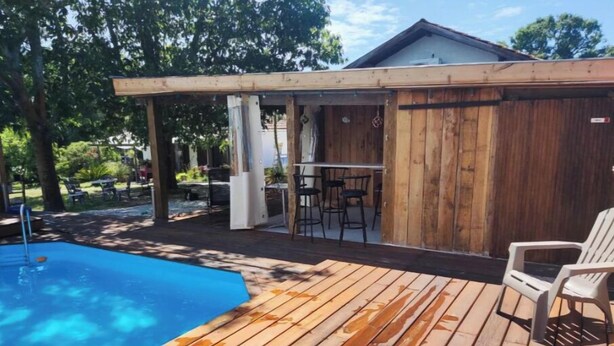 Una terraza de madera con piscina, mobiliario exterior y un área cubierta con sillas.