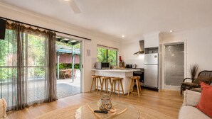 A modern kitchen with a central island, a dining area with wooden stools, a glass coffee table, and a view of a garden through large windows. - Cape Byron Retreat (Skinners Shoot)