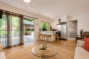 A modern kitchen with a central island, a dining area with wooden stools, a glass coffee table, and a view of a garden through large windows. - Cape Byron Retreat (Skinners Shoot)