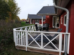A wooden deck with a white railing, a table with chairs, and a red building with a dark roof. - 4 Star Holiday Home in Grossenbrode (Grossenbrode)