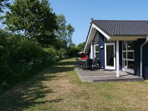 Ein modernes Haus mit einer Veranda und einem Essbereich mit Stühlen und einem Tisch, sowie einem großen Fenster.